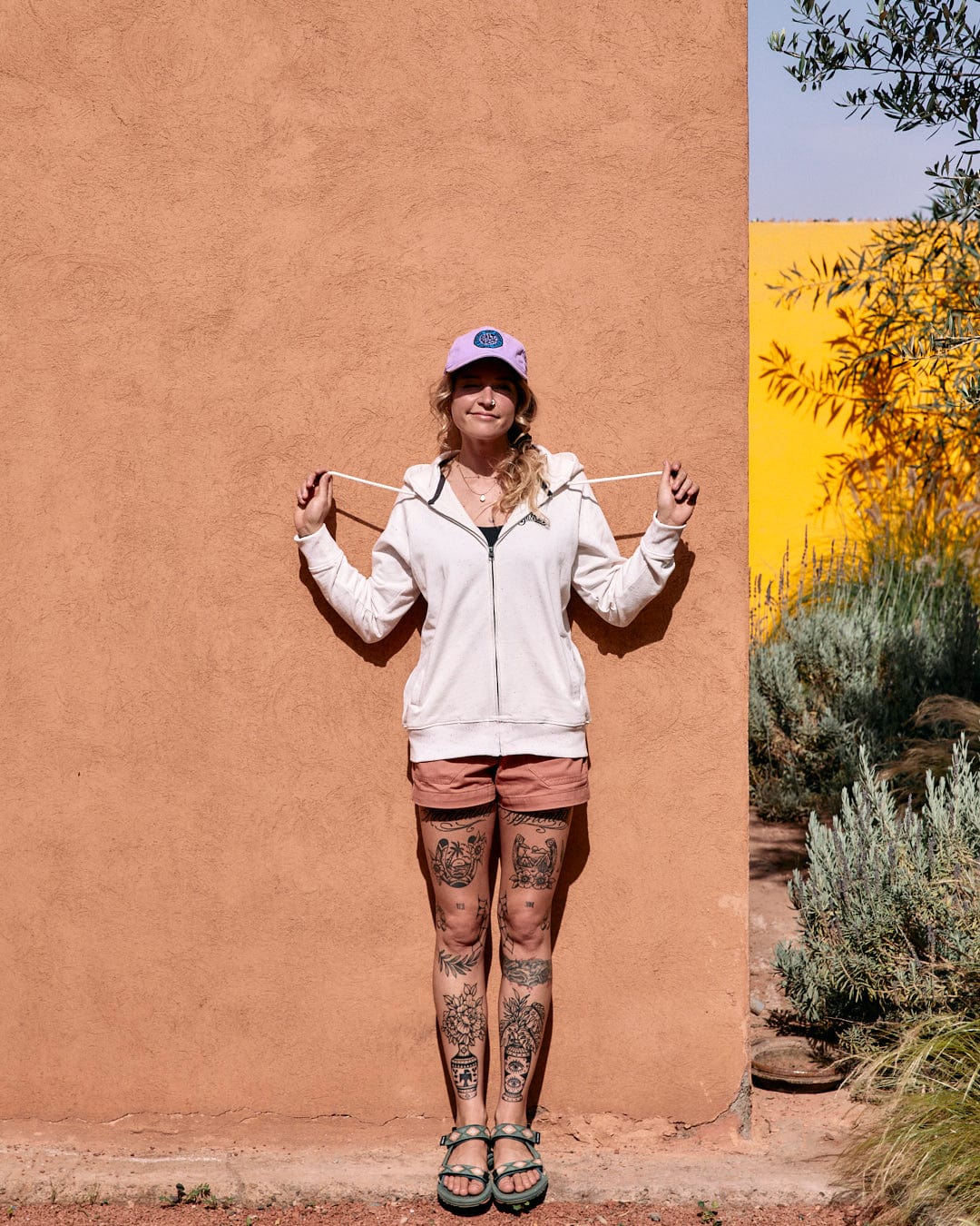 A person stands smiling in front of a pink wall, wearing a white Ginny - Womens Zip Hoodie - Cream with Saltrock branding, shorts, and sandals, and showing off tattoos on their legs.