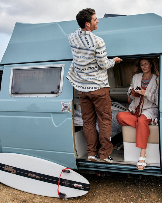 A man stands beside a camper van, adorned with Saltrock branding, looking at a woman seated inside, with a Godrevy 2 - Mens Cargo Trousers - Brown surfboard leaning against the vehicle.