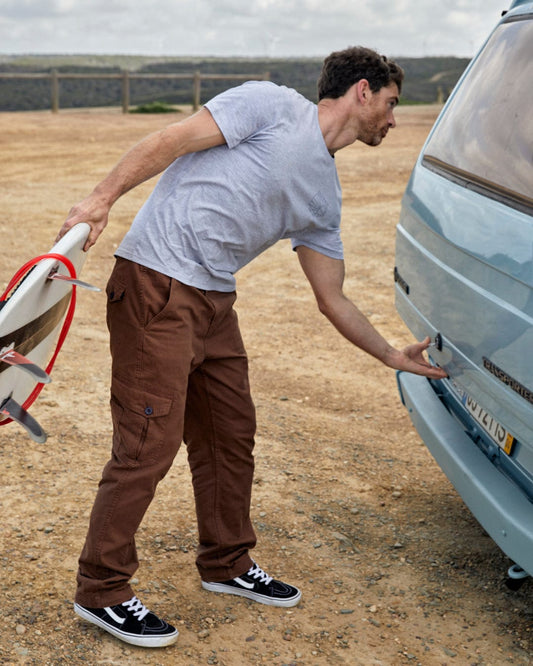 A man holding a surfboard checks the back of a vintage van, its Godrevy 2 - Mens Cargo Trousers - Brown by Saltrock adding a unique touch.