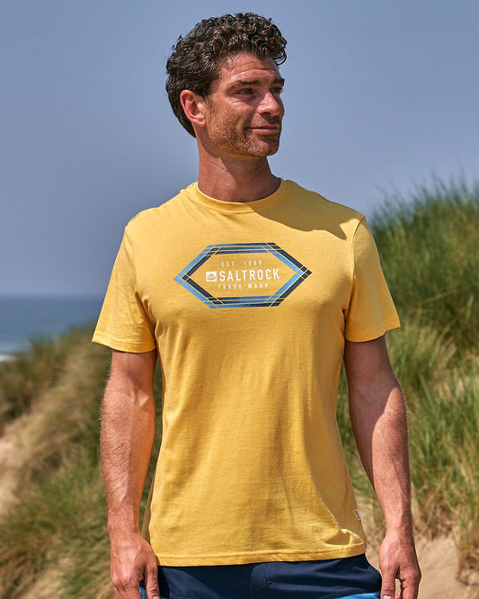 A man wearing a Saltrock Gradient Hex - Mens Short Sleeve T-Shirt - Yellow standing on a beach.