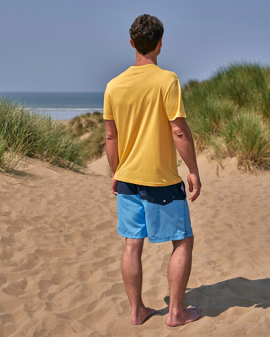 A man standing on a sand beach wearing a Saltrock Gradient Hex - Mens Short Sleeve T-Shirt - Yellow and blue shorts.