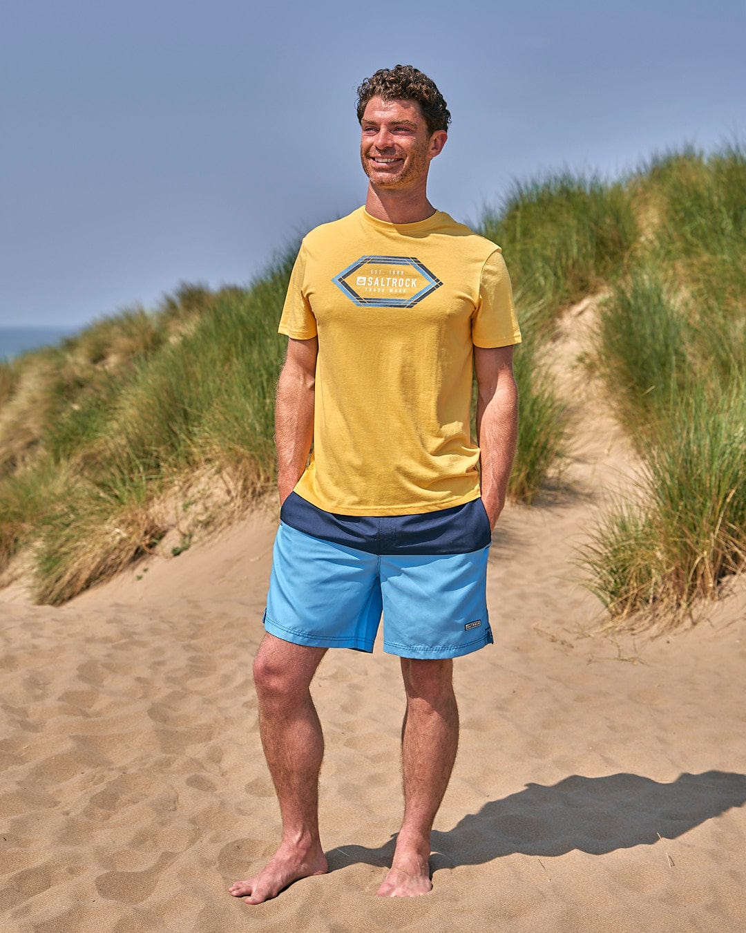 A man in a yellow Gradient Hex - Mens Short Sleeve T-Shirt - Yellow Saltrock standing on a sand dune.