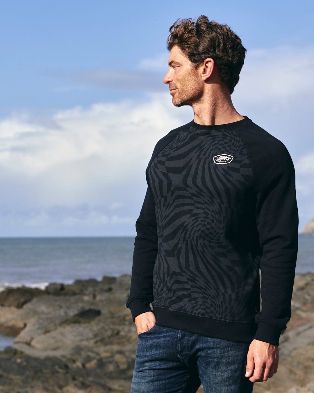 A man wearing a Grip It - Mens Crew Neck Sweat - Black sweatshirt with geometric print stands on a rocky beach, showcasing the Saltrock branding.