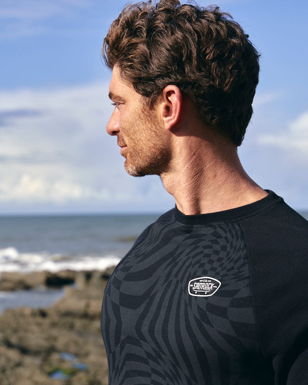 A man wearing a Saltrock Grip It - Mens Crew Neck Sweat - Black with geometric print, looking at the ocean.