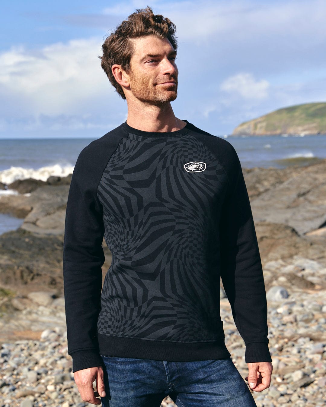 A man is standing on a beach wearing a black Saltrock sweatshirt featuring the Grip It logo.