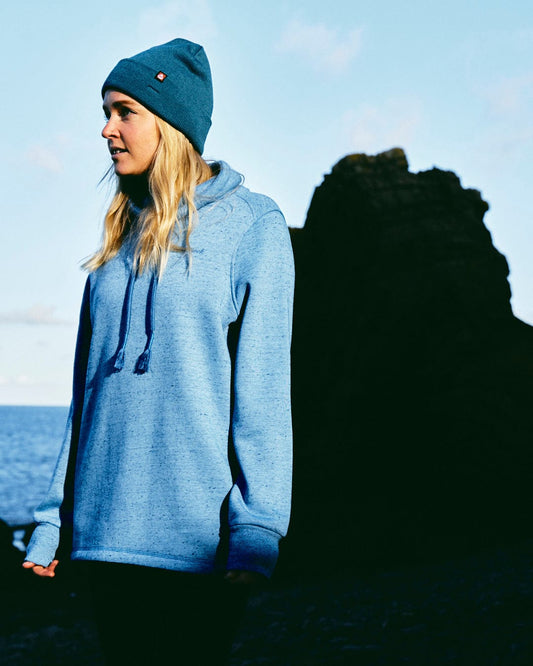 A chic woman, dressed in a comfortable Saltrock Harper - Womens Longline Pop Sweat - Light Blue and a beanie, stands in front of the serene ocean.