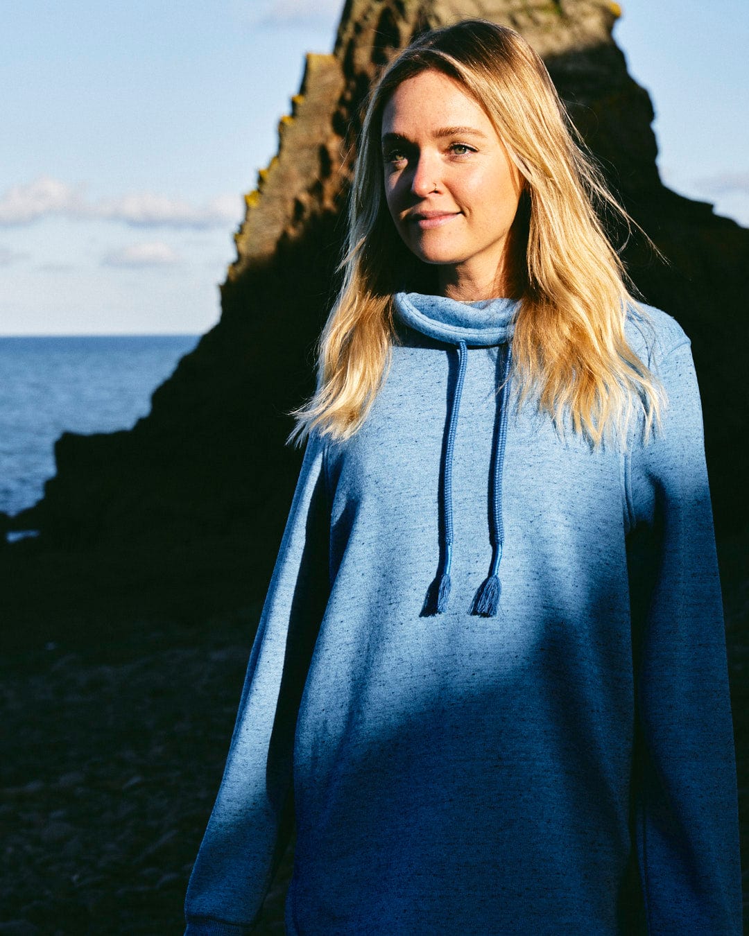 A chic woman in a Saltrock Harper - Womens Longline Pop Sweat - Light Blue hoodie standing by the ocean, exuding comfort.