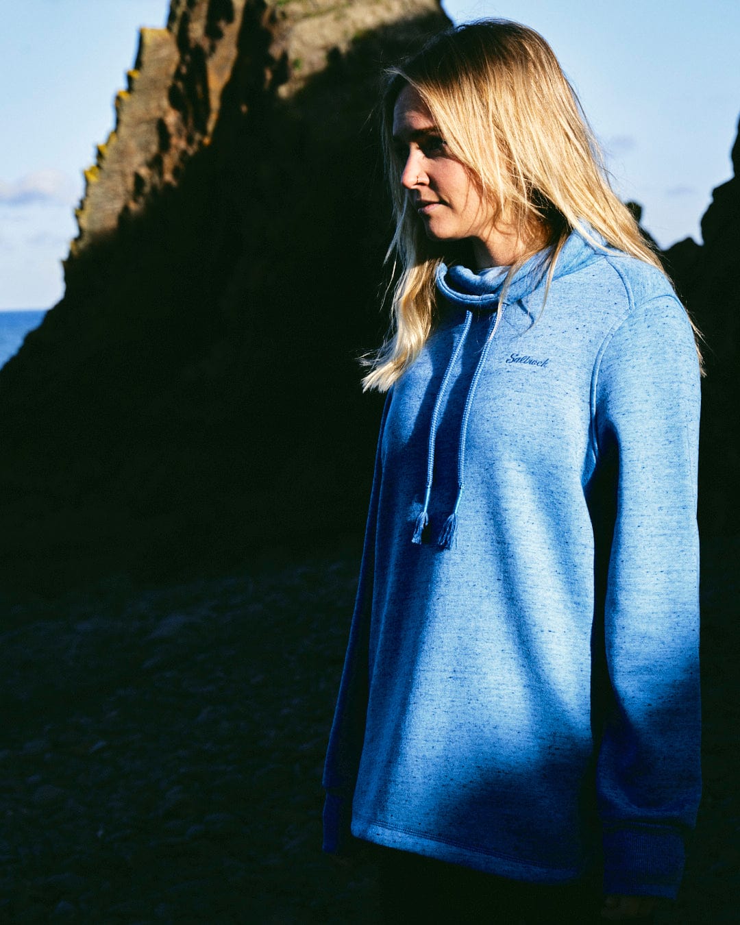 A chic woman wearing the Saltrock Harper - Womens Longline Pop Sweat - Light Blue hoodie standing in front of rocks.