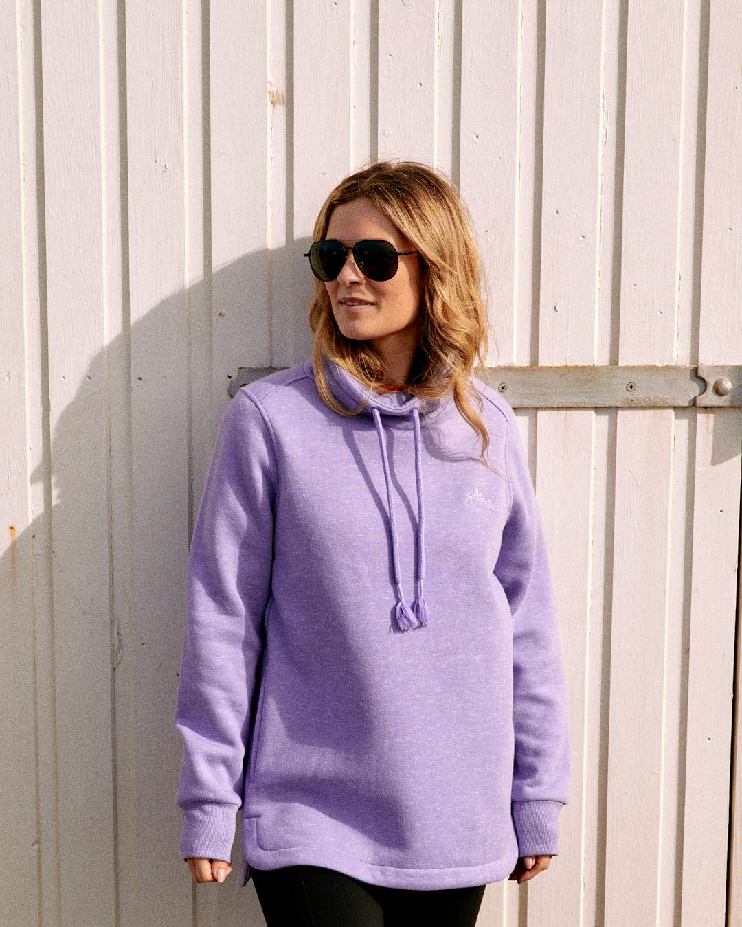A person wearing a Saltrock Harper Women's Longline Pop Sweat hoodie in lilac and sunglasses stands in front of a white wooden backdrop.