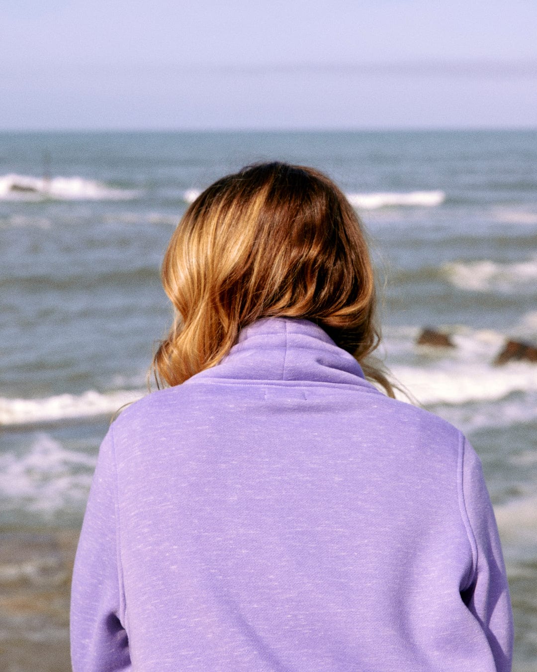 Person with long hair in the Saltrock Harper Womens Longline Pop Sweat in lilac, featuring embroidered branding, looking out at the ocean waves.