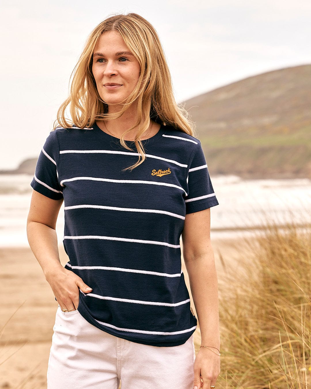 A woman wearing a Saltrock Hartland - Womens Striped Short Sleeve T-Shirt - Blue standing on the beach.