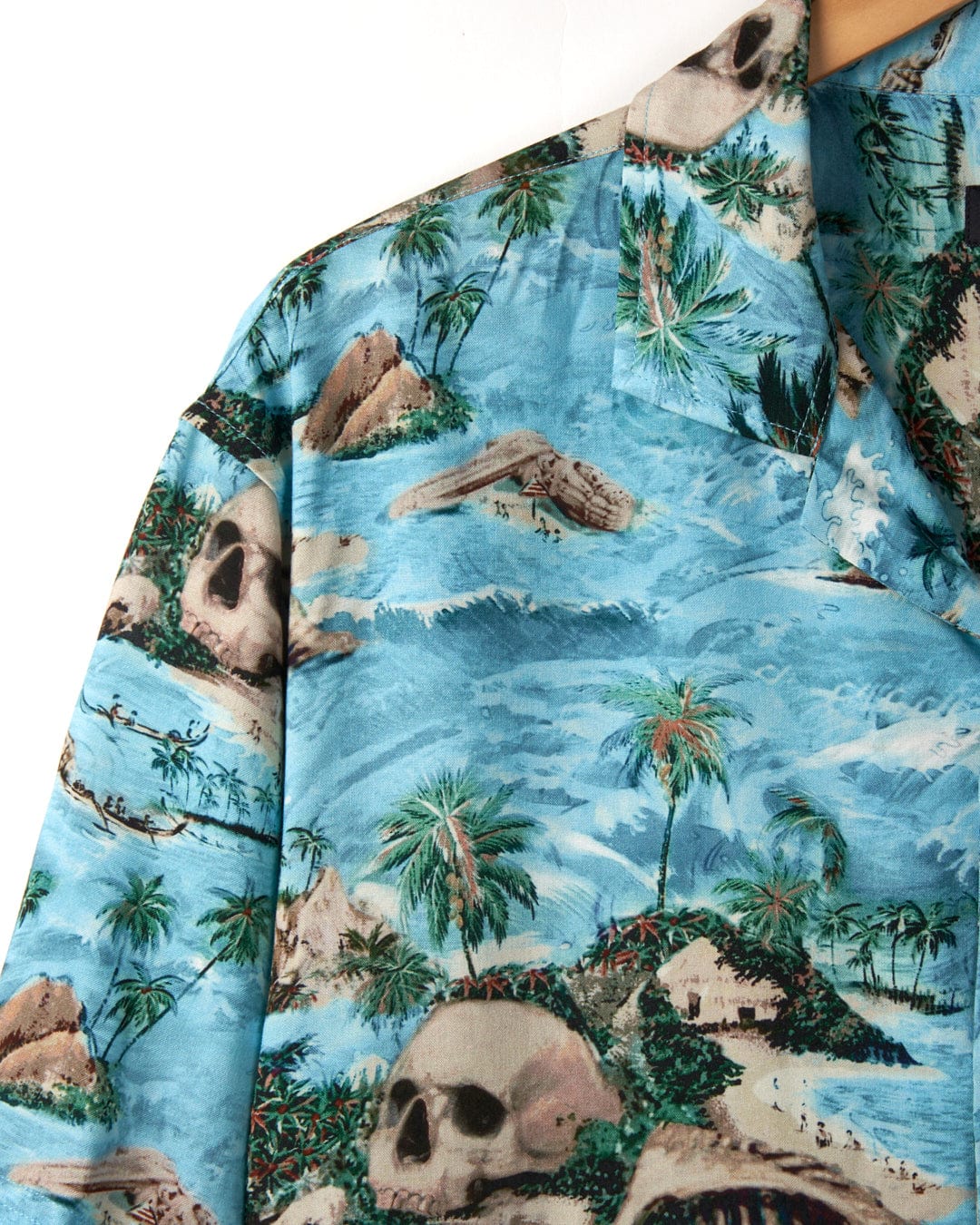 Close-up of a Saltrock Hawaiian Isle - Men's Short Sleeve Shirt - Blue featuring a skull island print with palm trees and ocean waves on a light blue background.