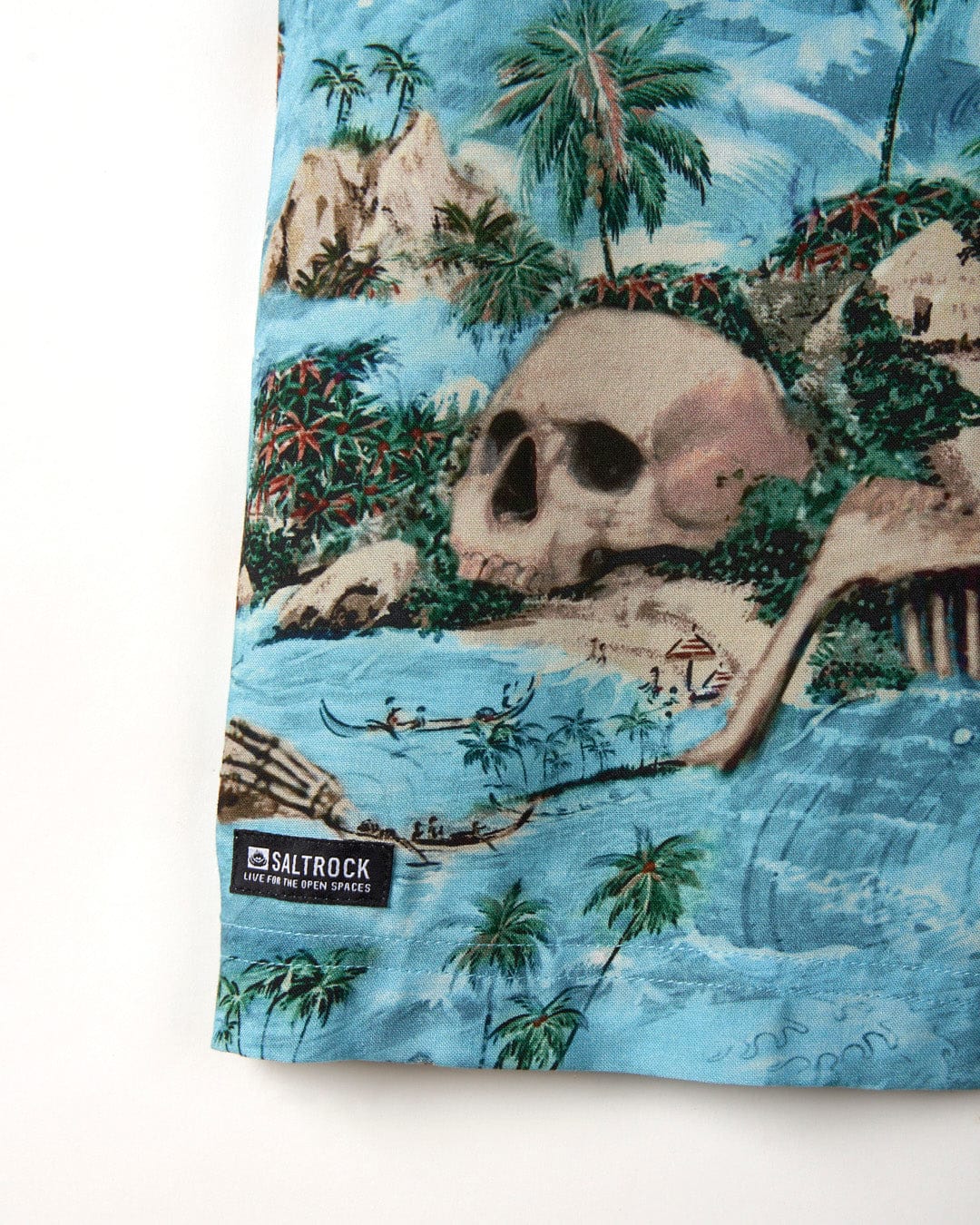 A close-up of a Hawaiian Isle - Mens Short Sleeve Shirt in Blue branded by Saltrock, featuring a tropical scene with palm trees, mountains, skull island print, and small figures on a blue background.