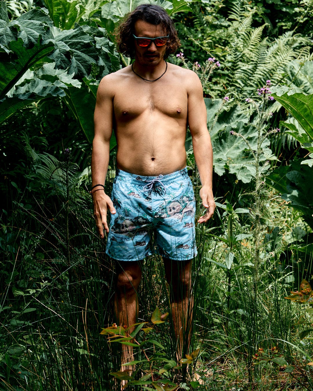 A shirtless man wearing sunglasses and Saltrock's Hawaiian Isle Men's Swimshorts in blue, featuring an elasticated waist, stands in tall grass surrounded by dense green foliage.