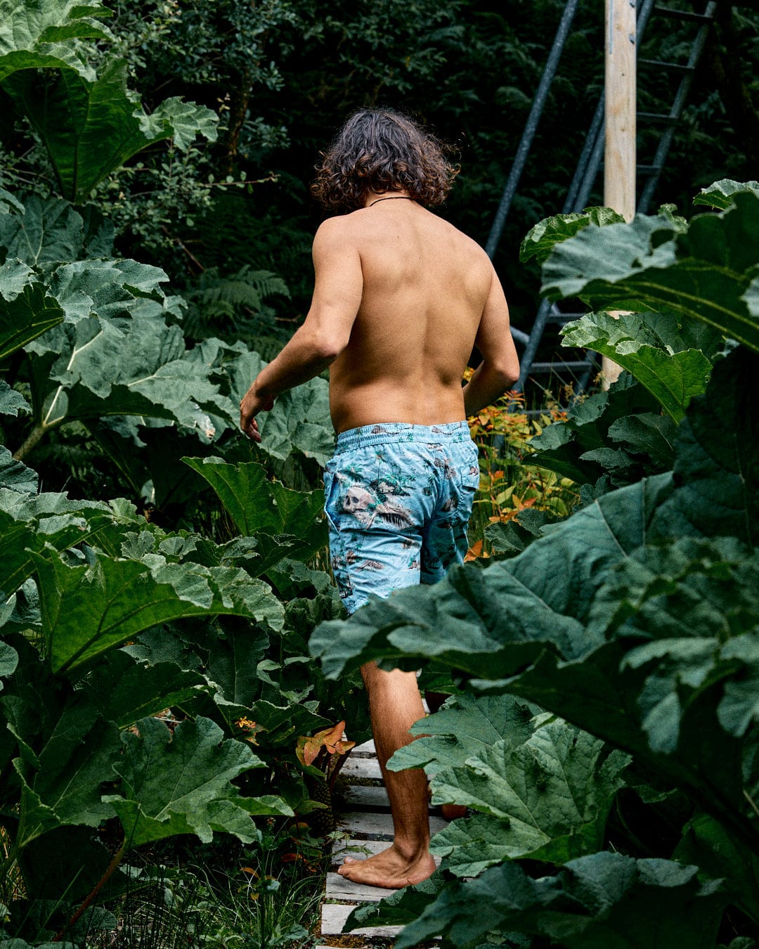 A barefoot, shirtless person walks along a narrow path lined with lush green plants, wearing Saltrock’s Hawaiian Isle Men’s Swimshorts in blue.
