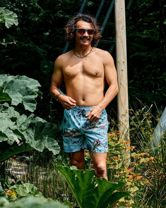 A man with curly hair and sunglasses stands shirtless among large green plants, wearing Saltrock's Hawaiian Isle Men's Swimshort in blue with an elasticated waist.