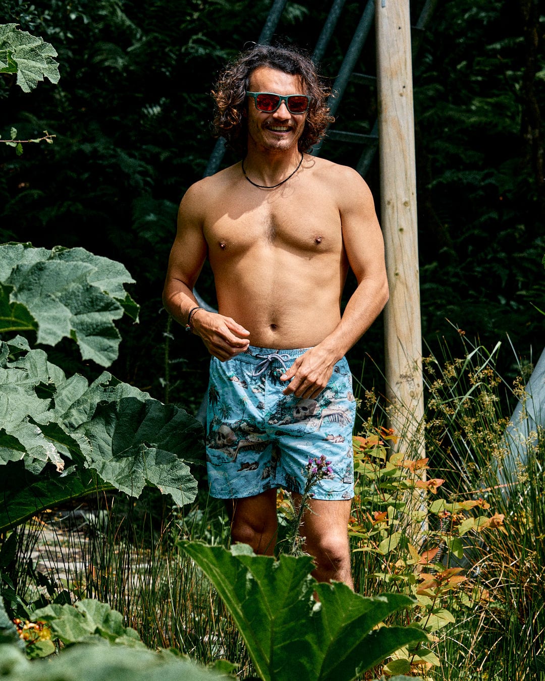 A man with curly hair and sunglasses stands shirtless among large green plants, wearing Saltrock's Hawaiian Isle Men's Swimshort in blue with an elasticated waist.