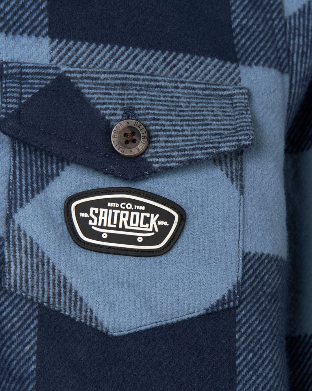 Close-up of a blue and grey plaid Hawkins Hooded Long Sleeve Check Shirt by Saltrock, with front chest pockets, featuring a branded patch and a button on one of the pockets.