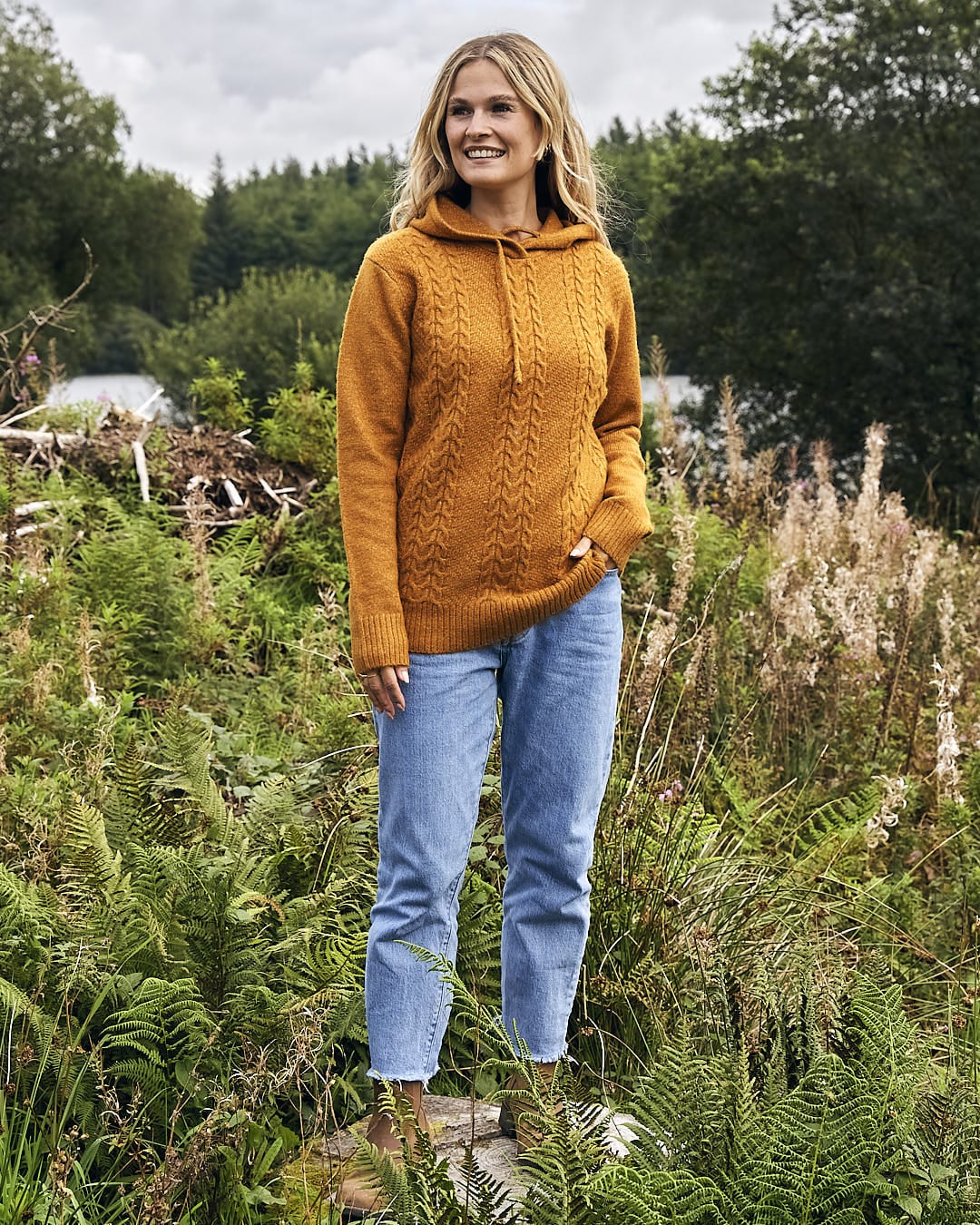 A woman standing in a field wearing a Saltrock mustard hoodie and jeans.