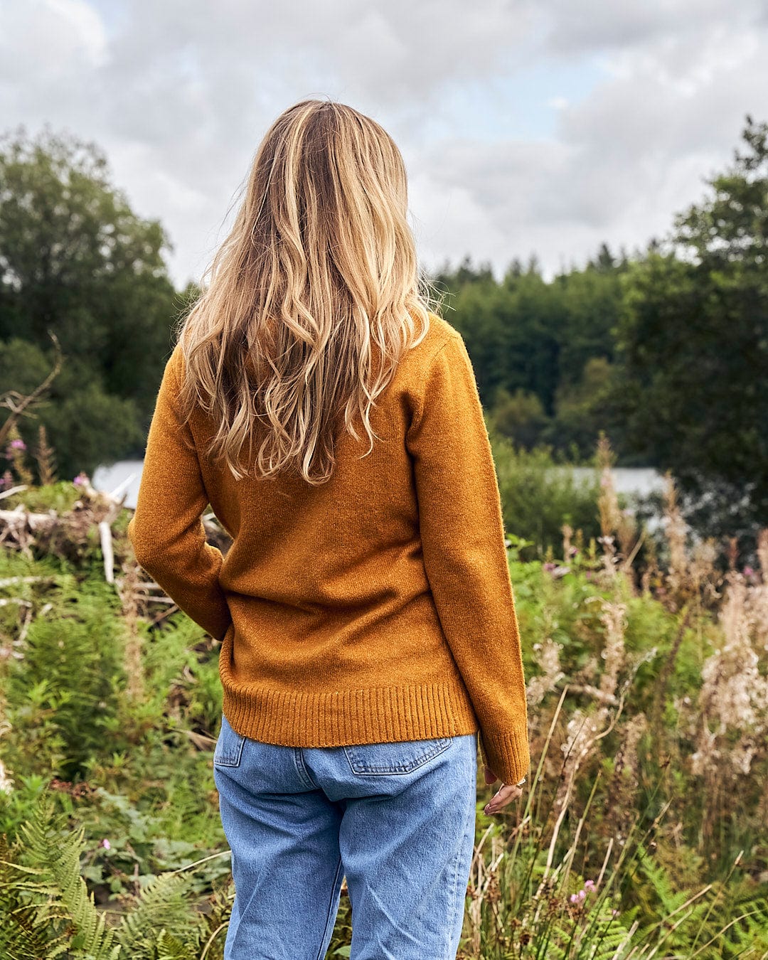 A woman standing in a field wearing a Saltrock Hele Bay 2 - Womens Knitted Jumper - Yellow sweater and jeans.