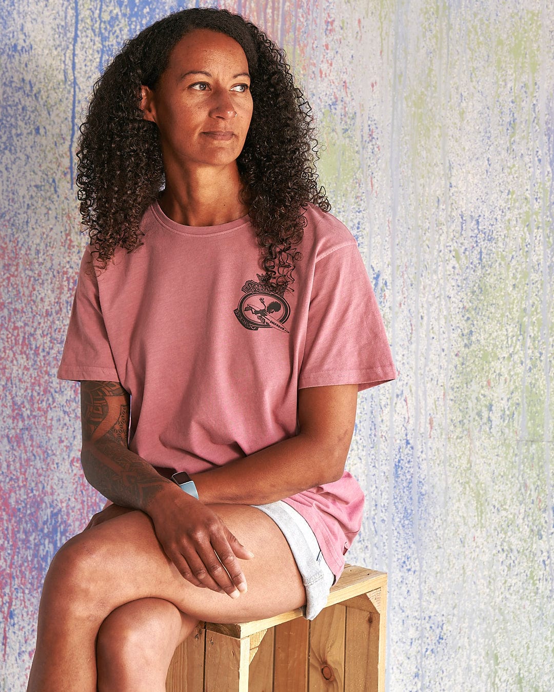 A woman wearing a Saltrock Heraldic Tok - Limited Edition 35 Years T-Shirt sitting on a wooden box.
