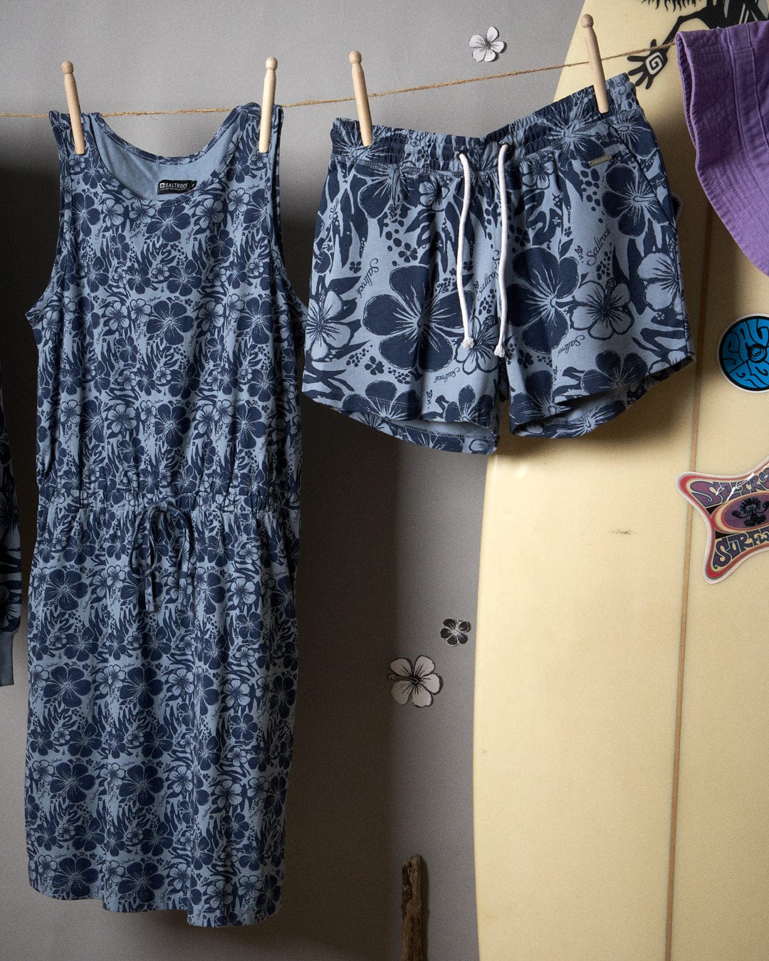 Hibiscus Bauhaus - Womens Midi Dress - Blue dress and shorts hanging on clothespins against a wall, with a surfboard and Hawaiian hibiscus stickers visible on the right side.