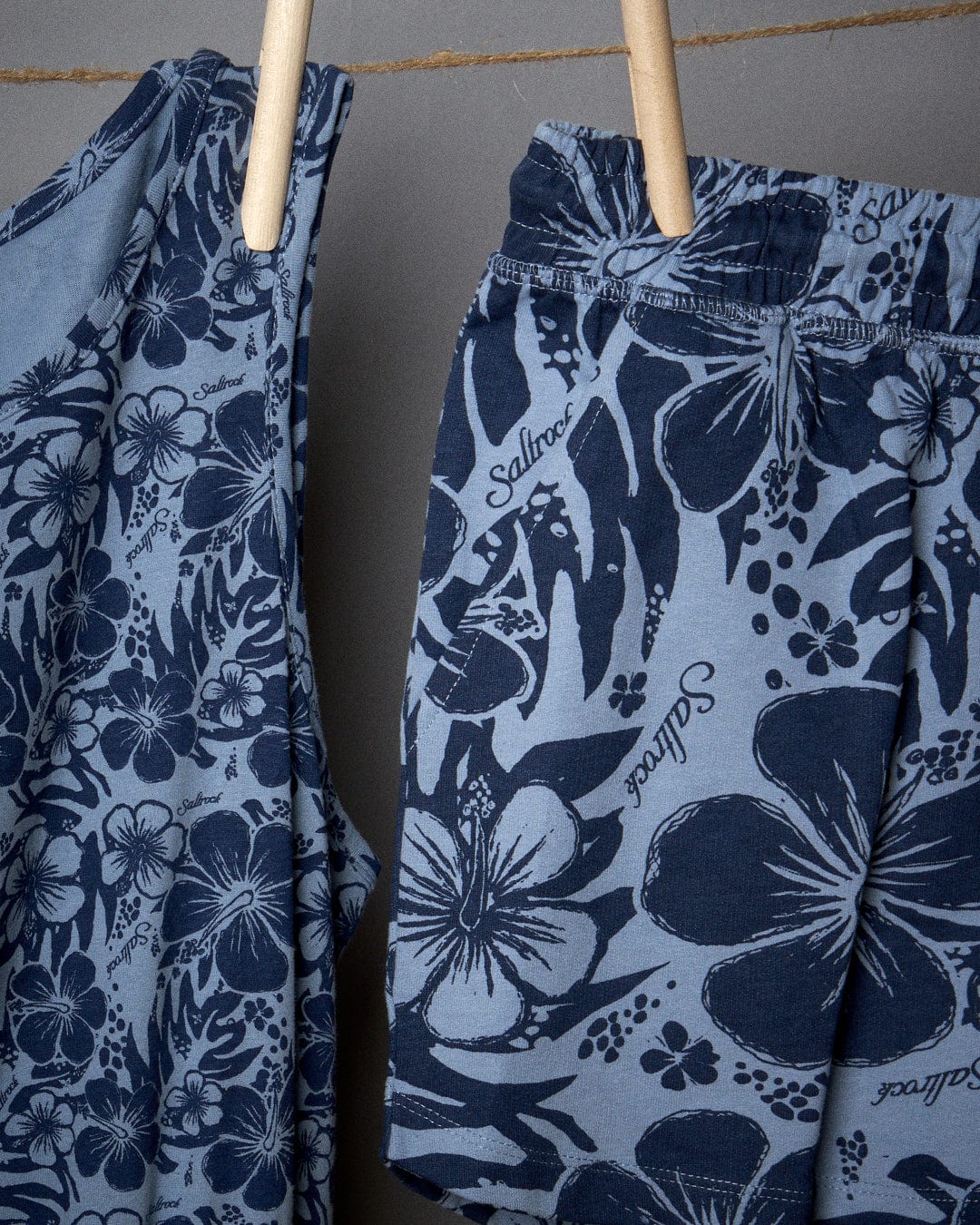 Two pairs of Saltrock branded swim shorts with a Hibiscus Bauhaus pattern hanging on wooden hangers against a gray background.