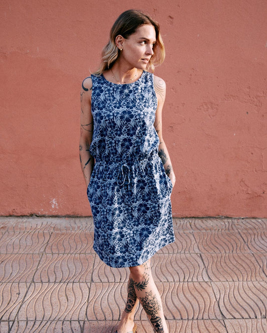 Woman in a blue Saltrock Hibiscus Bauhaus Womens Midi Dress with tattoos on her arms and legs, standing against a pink wall, looking away thoughtfully.