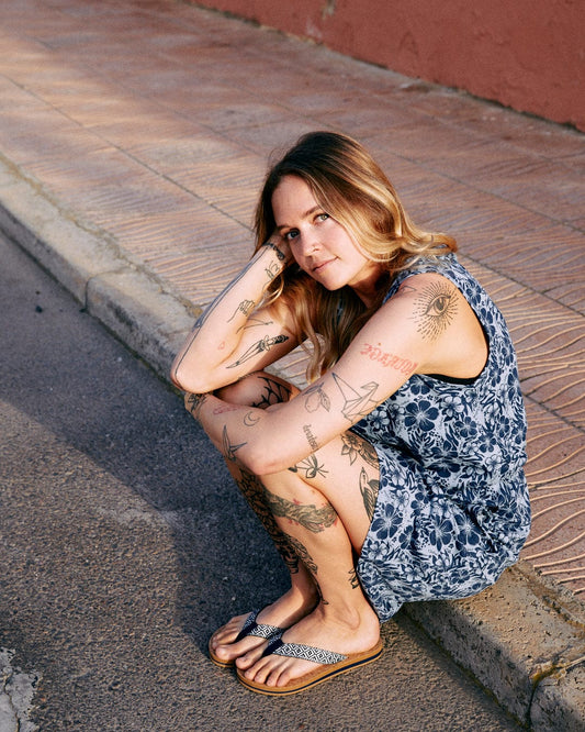 Woman with tattoos sitting on a sidewalk, wearing a blue Saltrock Hibiscus Bauhaus midi dress and flip-flops, looking thoughtfully at the camera.