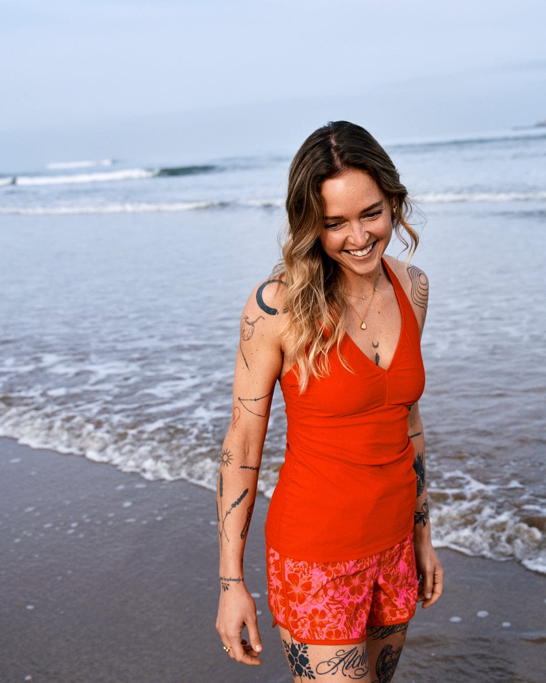 A woman in a red floral print top smiles as she walks along a beach, with waves in the background and tattoos visible on her arms wearing Saltrock's Hibiscus Womens Boardshorts in Red/Pink.