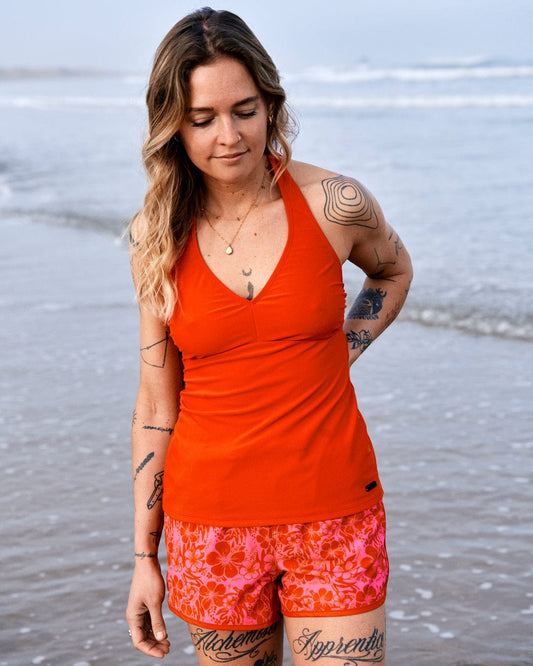 A woman in an orange tank top and Saltrock Hibiscus Women's Boardshorts in Red/Pink stands on a beach, looking downwards, with visible tattoos on her arms.
