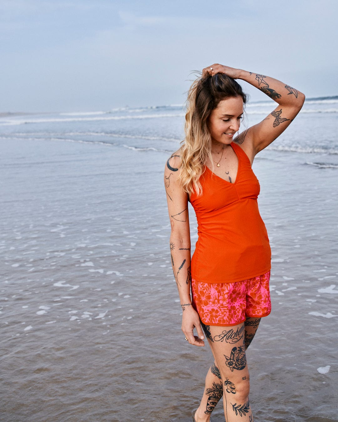 A woman with tattoos standing on a beach, smiling and adjusting her hair, wearing an orange top made of Repreve recycled material and Saltrock's Hibiscus - Womens Boardshorts - Red/Pink.