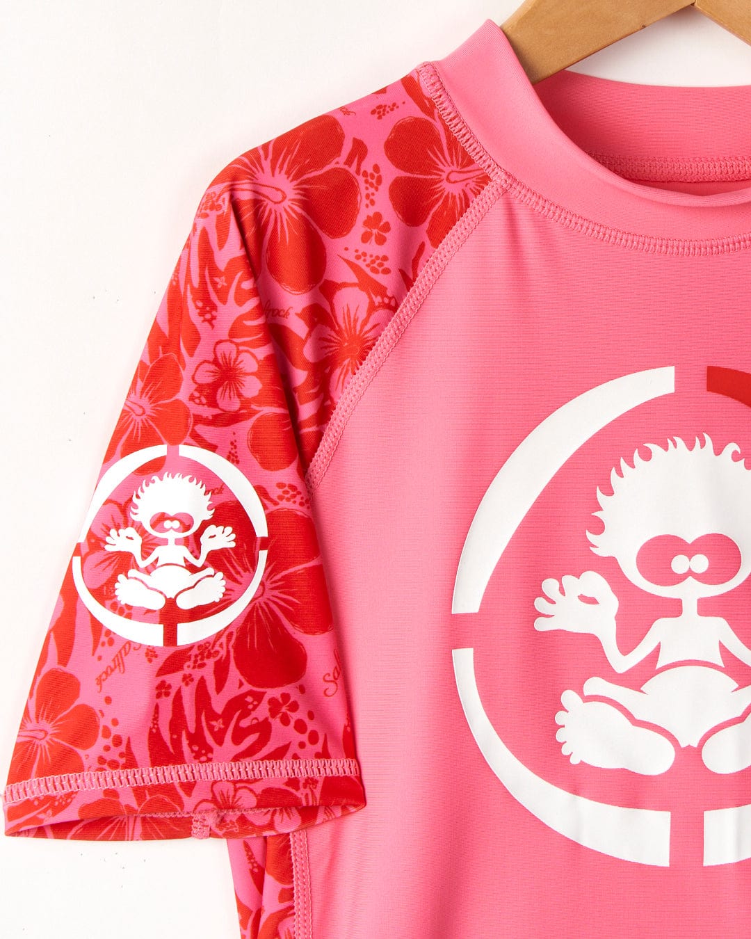 A close-up of a pink Saltrock Hibiscus rash vest with a graphic of a cartoon monkey in the center, hanging on a wooden hanger against a white background.
