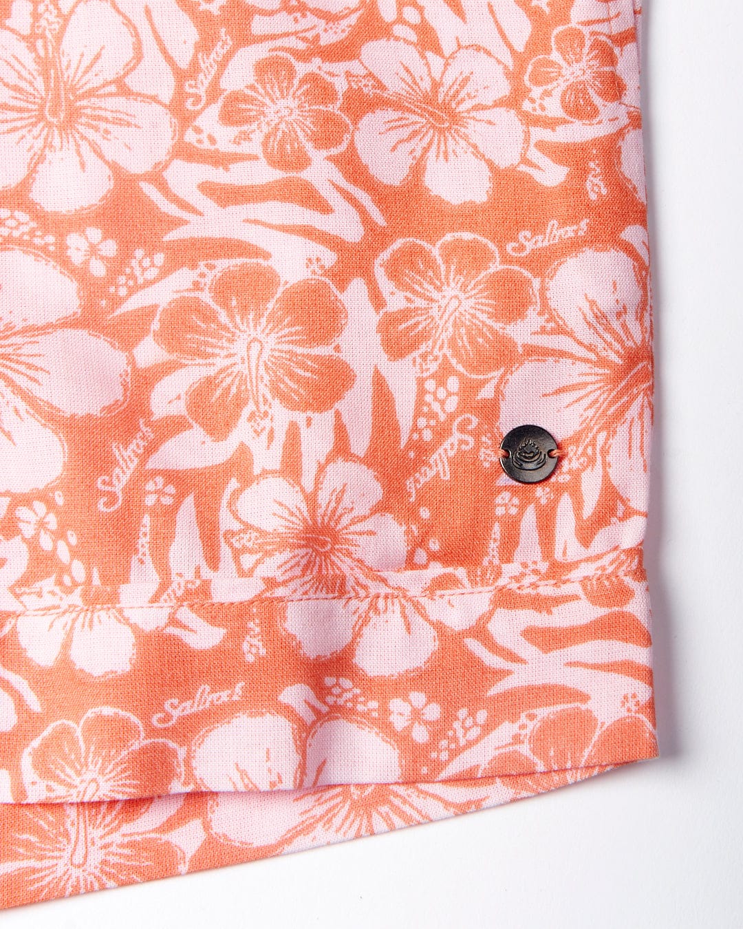 Close-up of a fabric with a pink and white hibiscus floral pattern featuring the Saltrock logo, and a black button on a cuff.