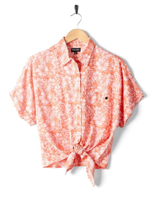 A coral-colored Saltrock Hawaiian shirt with hibiscus floral print, hanging on a hook against a white background.