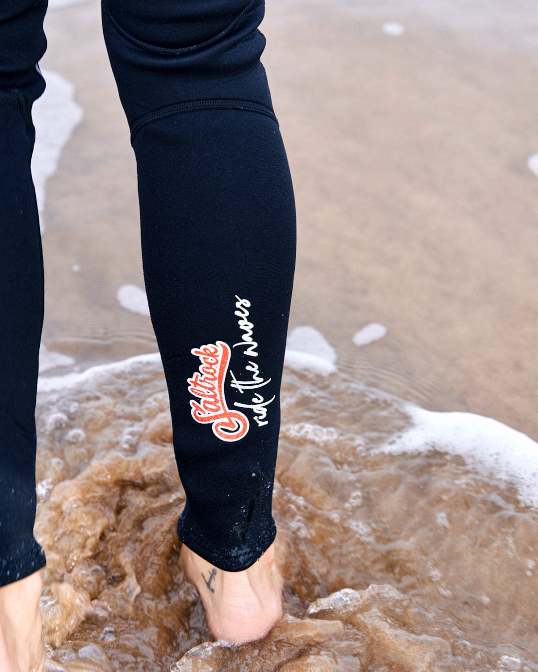 Close-up of a person's lower legs in black neoprene Saltrock wetsuit with "champion de la mer" written on them, standing in shallow ocean water.