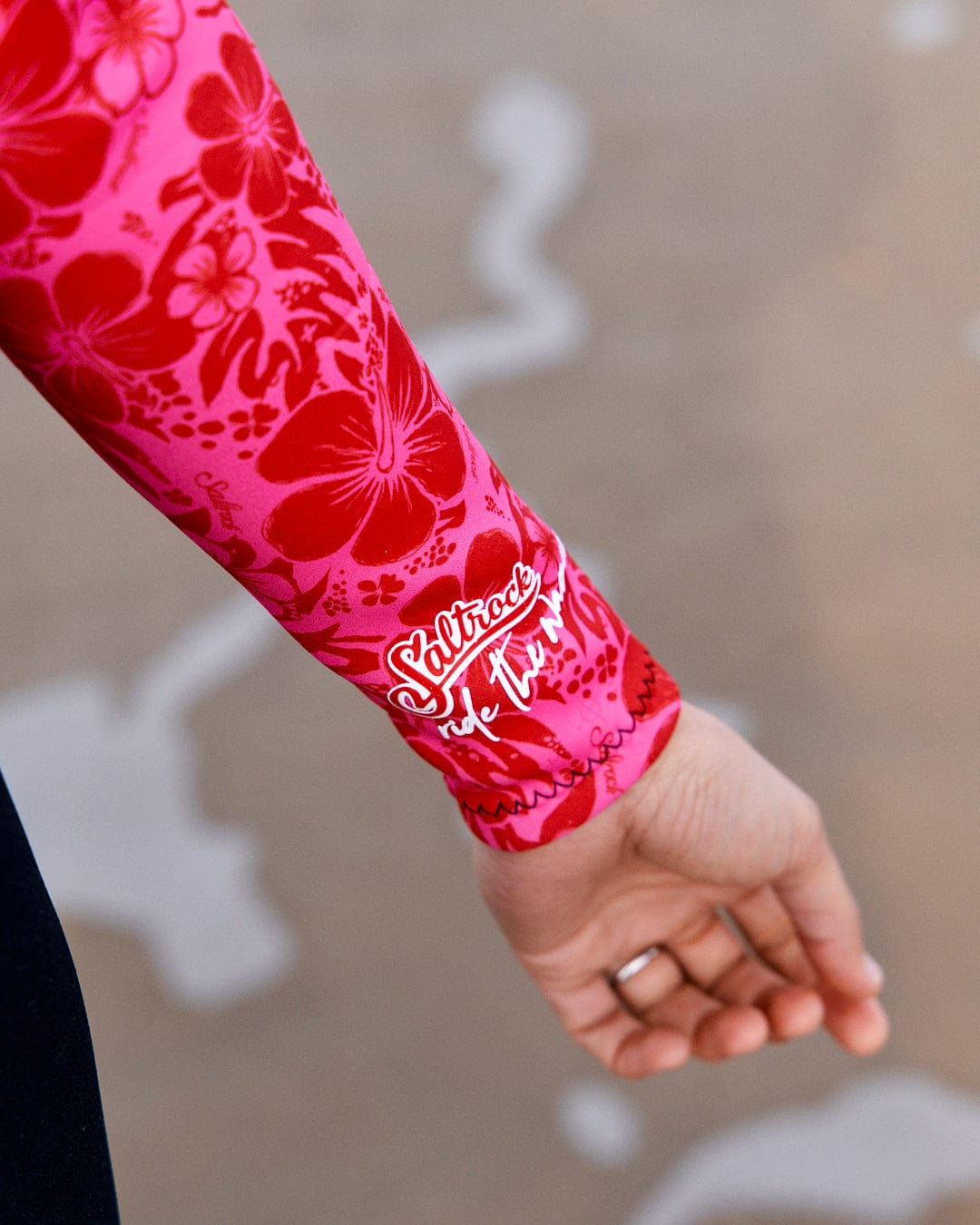 Close-up of an arm wearing a bright red neoprene wetsuit sleeve with Saltrock Hibiscus - Womens Full Wetsuit - Pink printed on it, against a sandy background.