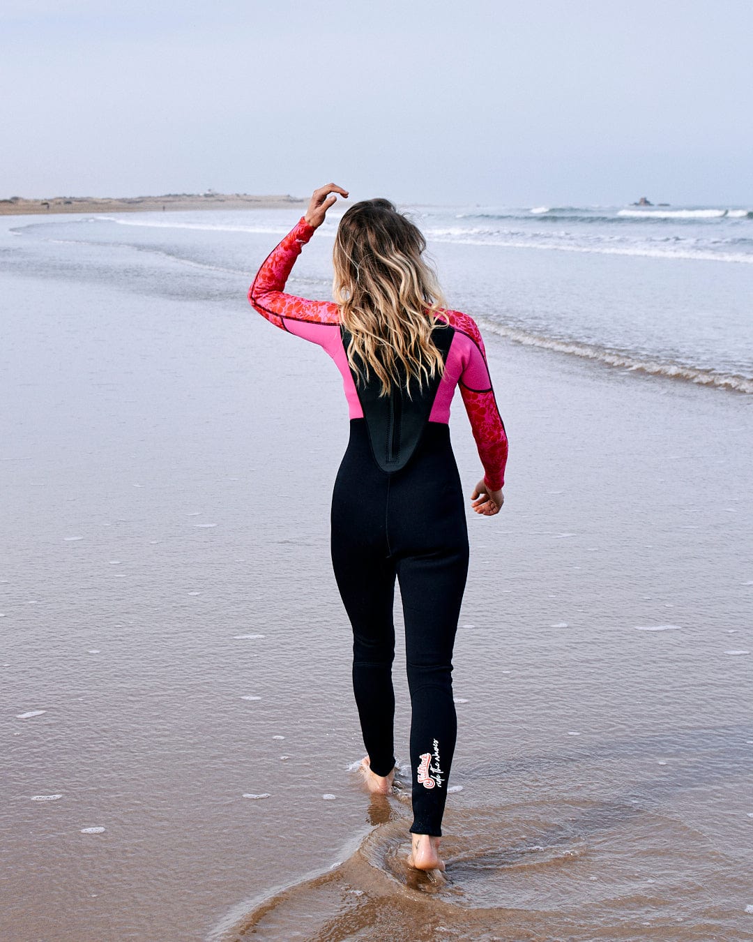 A woman in a Saltrock neoprene Hibiscus Womens Full Wetsuit in Pink walks along the beach, looking towards the sea, with her hand shielding her eyes.