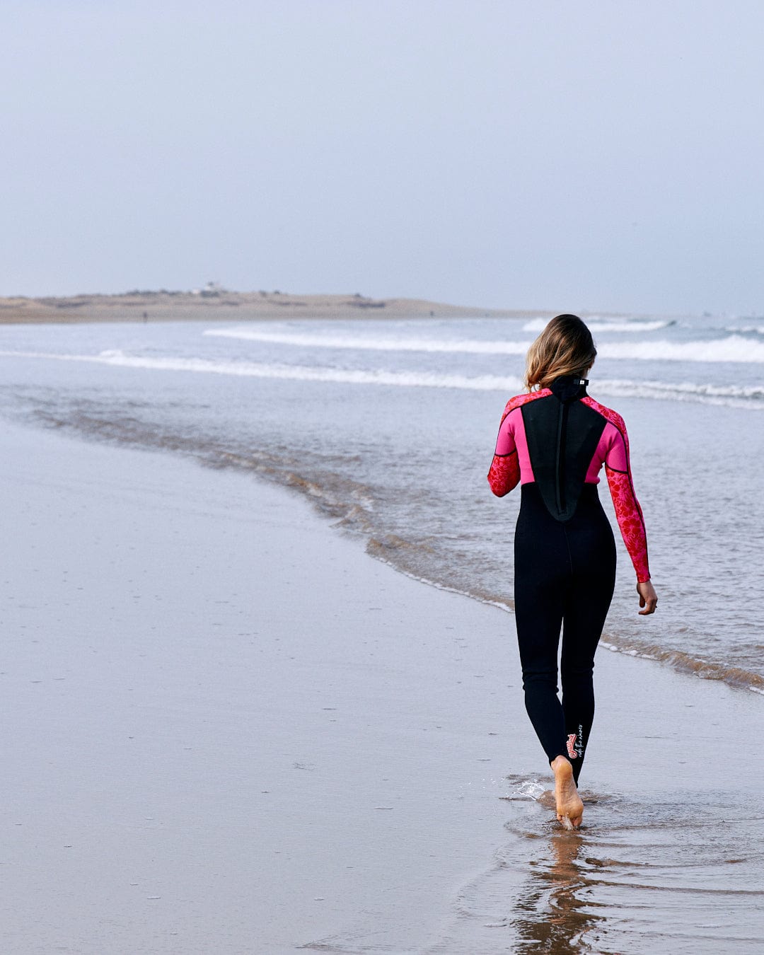 A woman in a Saltrock neoprene wetsuit walks along the shoreline, her back to the camera, facing the waves on a cloudy day.