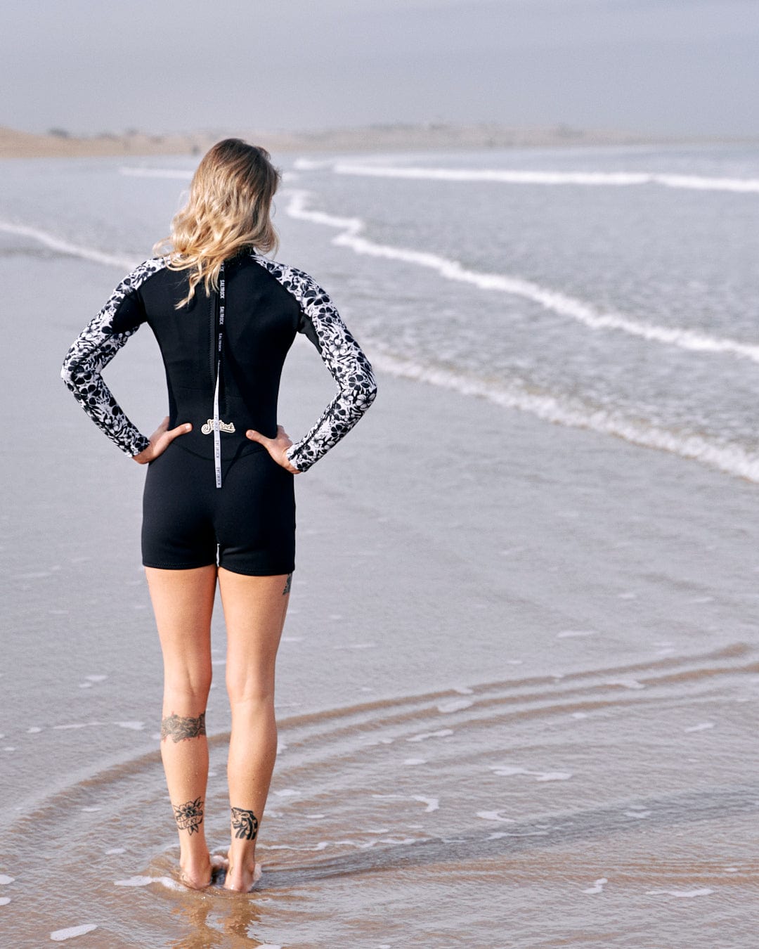 Woman in a Hibiscus - Womens Shortie Wetsuit - Black neoprene wetsuit by Saltrock standing at the shore, looking at the sea, with her back to the camera.