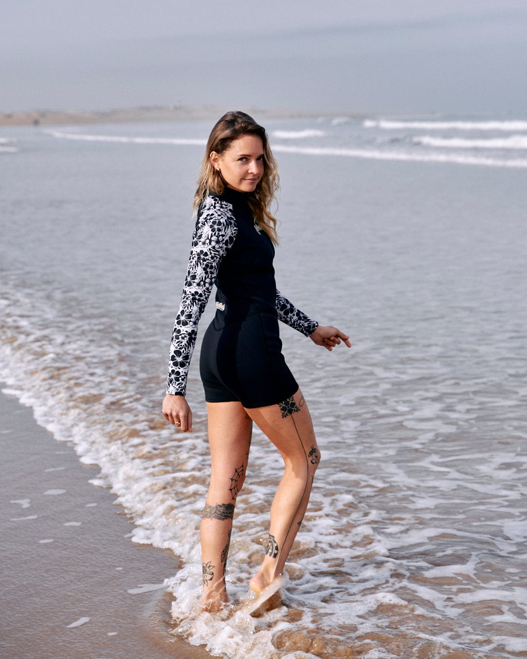 A woman in a Hibiscus - Womens Shortie Wetsuit - Black by Saltrock standing on a sandy beach with gentle waves at her feet.