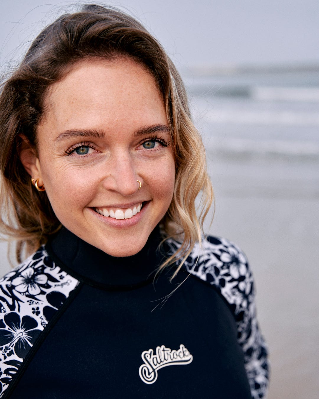 A smiling woman with blue eyes and a nose ring, wearing a Hibiscus - Womens Shortie Wetsuit in black with the brand "Saltrock" on the beach.