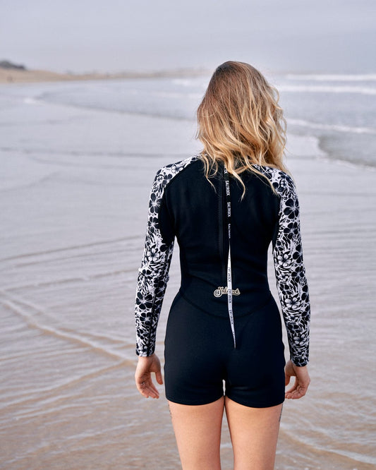 Woman in a Saltrock Hibiscus - Womens Shortie Wetsuit - Black neoprene wetsuit standing on a beach, looking at the ocean waves, with her back to the camera.