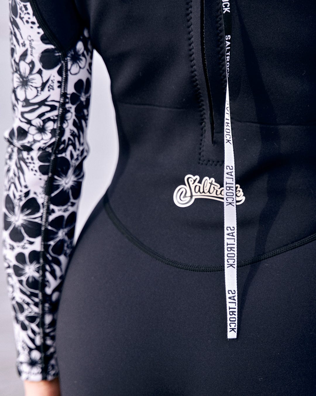 A close-up of a person wearing a black and white floral patterned sleeve and a neoprene wetsuit with a Saltrock logo and zipper pull detail, Hibiscus - Womens Shortie Wetsuit - Black.