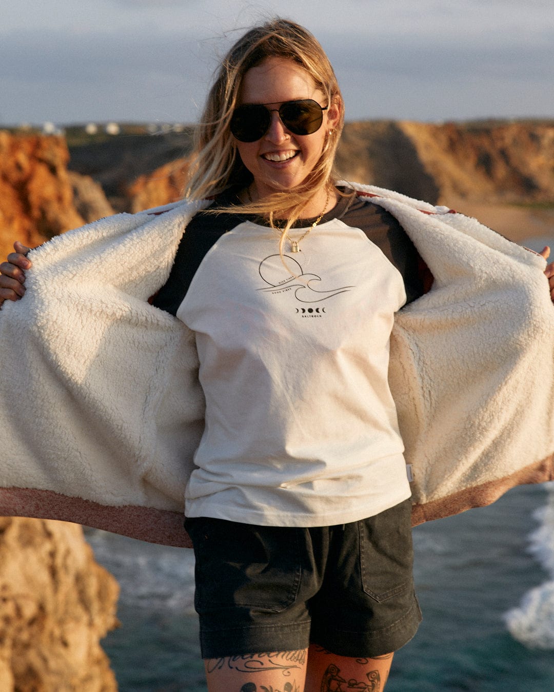 A woman in a Saltrock High Tides - Womens Raglan Long Sleeve T-Shirt - White and shorts is standing on a rock by the ocean.