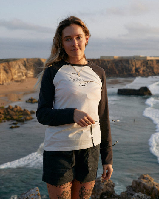 A woman with tattoos standing on a rock beside the ocean, her sleeves rolled up to reveal intricate designs in her Saltrock High Tides - Womens Raglan Long Sleeve T-Shirt - White.