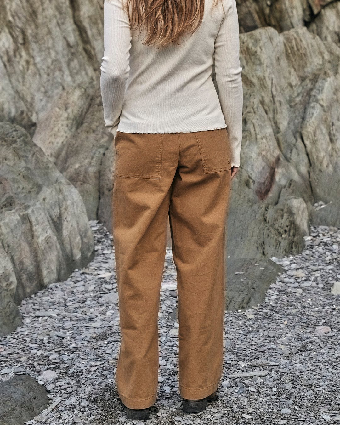 A woman is standing on a rocky beach wearing Saltrock's Hilda Twill - Womens Trouser - Light Brown for ultimate comfort.