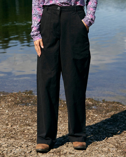 A woman is standing by a body of water wearing the Hilda Twill - Womens Trouser - Black by Saltrock.