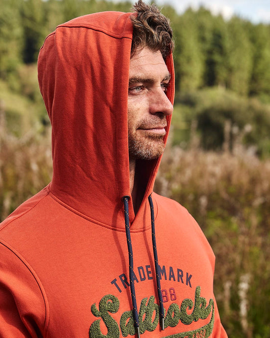 A man wearing an orange Home Run Chenille - Mens Pop Hoodie, with Saltrock embroidery, standing in a field.