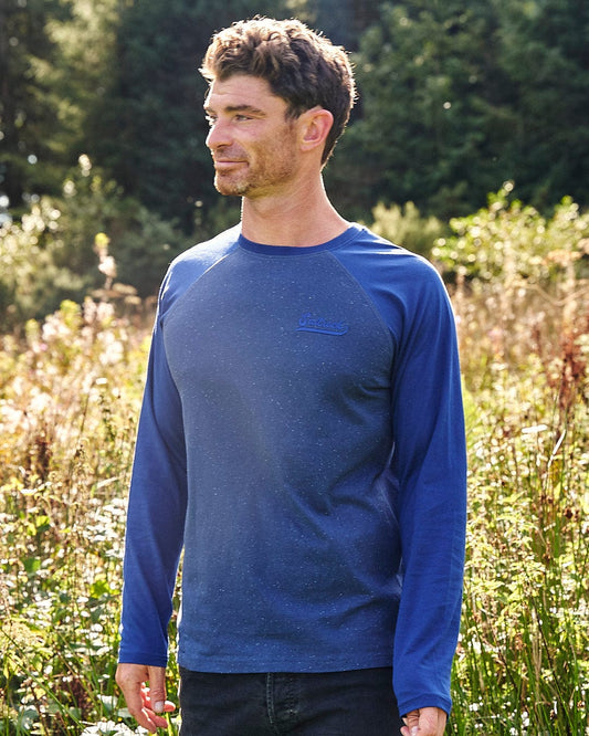 A man standing in a field wearing a blue Home Run Nep - Mens Raglan Long Sleeve T-Shirt from Saltrock branding.